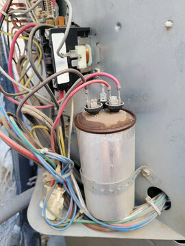 A rusty silver metal dual motor run capacitor connected to multiple colorful electrical wires inside an outdoor HVAC system compressor housing