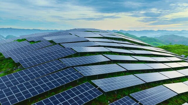 Rows of photovoltaic solar panels cover a grassy hillside beneath a cloudy blue sky in soft daylight, overlooking distant mountains.