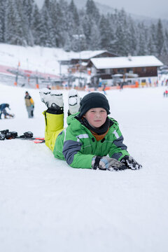 A boy lies on the snow during a ski lesson at a winter resort. The scene represents childhood learning, first skiing attempts, and winter sports practice.