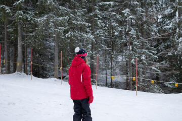A young girl stands on skis and looks across a snowy slope surrounded by forest at a winter resort. The scene represents youth skiing, winter recreation, and outdoor adventure. © Liudmila