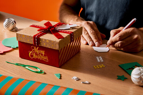 Man Writing on a Gift Tag Beside a Decorated Christmas Present Box on a Wooden Table