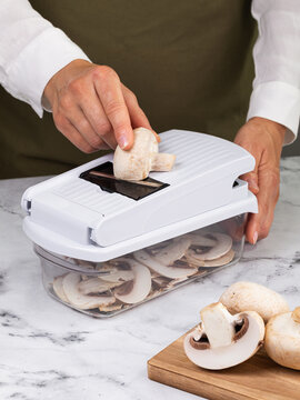 Woman slicing fresh button mushrooms into thin slices with manual mandoline slicer and container on marble countertop