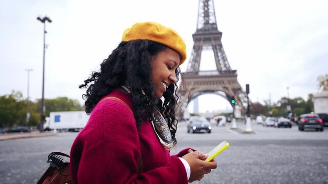 A smiling Black woman walks beside the Eiffel Tower while typing and reading messages on her yellow smartphone.