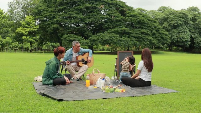 Asian kids painting in park. Asian family enjoy outdoor picnic with food and drinks in park sunny day, outdoor activitie with daughter father, mother grandfather. GrandFather playing acoustic guitar