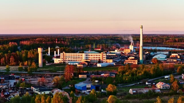 Aerial view of an industrial factory complex featuring a tall chimney and a water tower, surrounded by lush green forests during the golden hour in Latvia.
