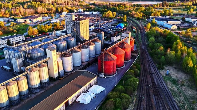 Aerial view of a grain silo facility with large metal and red cylindrical storage tanks, railway tracks, and surrounding greenery in Latvija.
