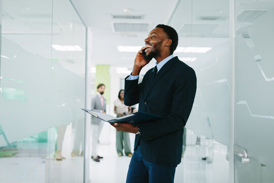 Black businessman laughing and talking on smartphone while holding documents. Standing in modern office corridor with diverse colleagues in background