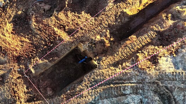 Aerial view of an excavation trench with visible tire tracks from heavy machinery and red and white safety tape marking the perimeter in Latvija.