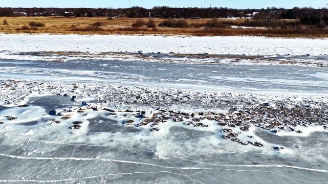 Aerial view of a frozen river surface with cracked ice patterns and snow-covered banks with distant trees in Latvia.