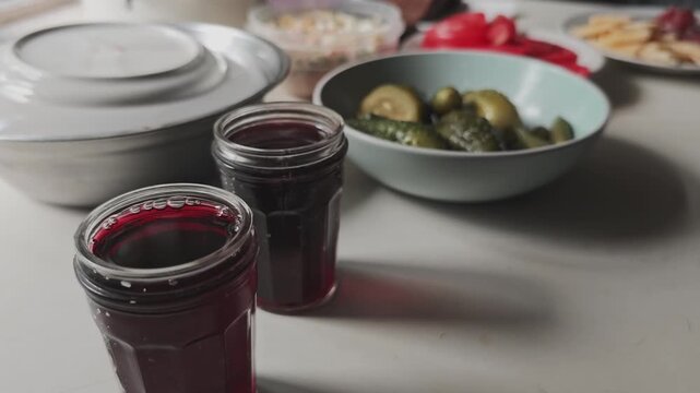 Rustic dinner table setup with various dishes, including pickled cucumbers, salads, and dark berry juice or wine in glasses for a family meal