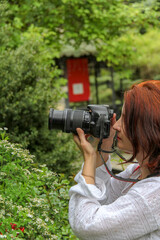 woman with red hair holds black digital camera near eye, profile, portrait, close-up, mirrorless, middle age, lens mounted, greenery in background, green plants, beautiful, romance, white blouse,  © Studio  GULL