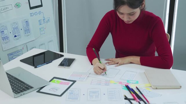 A focused young female designer in a red top sketches mobile app wireframes on paper while sitting at a white desk surrounded by a laptop tablet phone and colorful sketches in a bright modern studio