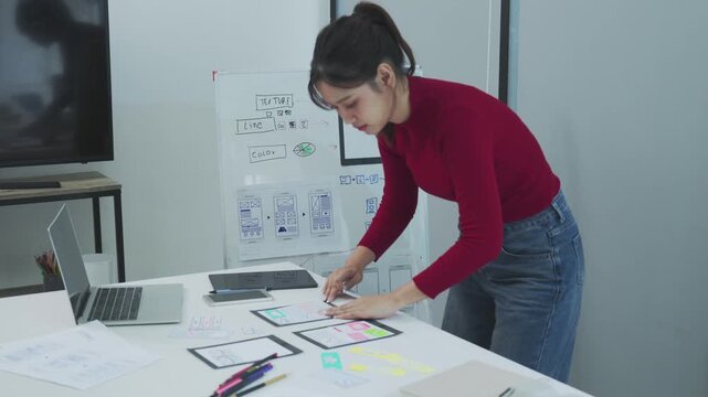 A young female designer in a red sweater arranges paper wireframes and sketches on a white table while working near a laptop and whiteboard in a bright modern studio with focused creative energy