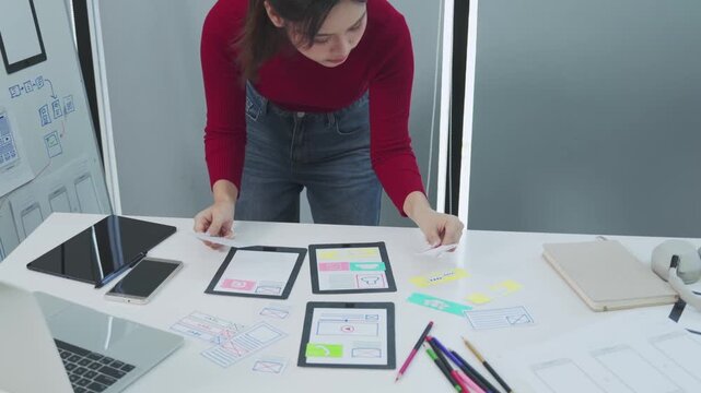 A young female designer stands at a white desk arranging paper wireframes and app interface cards while planning a mobile application layout with a tablet phone and sketches nearby in a bright modern 