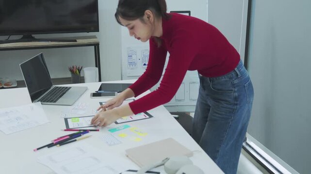 A young female designer in a red top leans over a bright office desk arranging colorful wireframe sketches and mobile app prototypes while a laptop and phone sit nearby in a creative workspace with wi