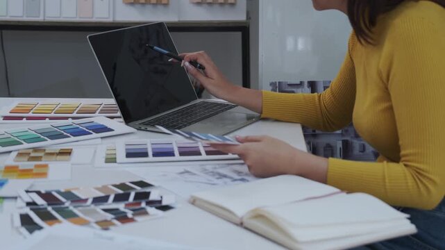 Young woman in a yellow sweater works at a bright studio desk while holding a fan of paint color swatches and a pen as she compares palette samples with her laptop and open sketchbook in a creative in