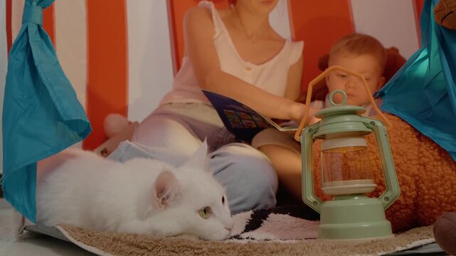 A white cat lies peacefully by a woman and her son as she reads a storybook inside a colorful tent, creating a warm and cozy atmosphere for family bonding.