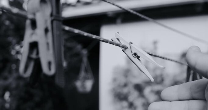 Black and white photo of three clothespins hanging on a wire clothesline outdoors. Soft blurred background with garden plants and a fence. Vintage film grain style. Everyday rustic scene.