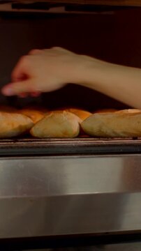 close eye-level kitchen view of hand reaching into warm oven to adjust row of golden bread rolls on metal tray showing domed glossy crusts in warm glow now just