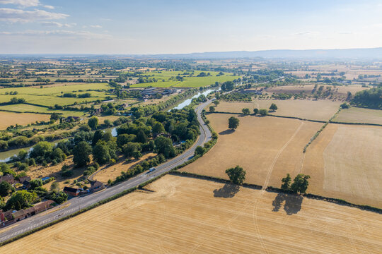 Aerial view of a winding road cutting through golden agricultural fields and lush green trees alongside a calm river in the rural landscape of Cheltenham, England, United Kingdom.