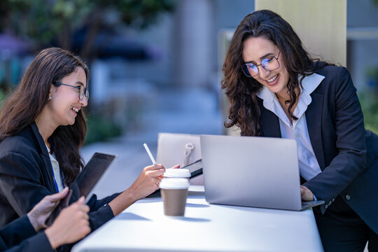 A diverse team of three Hispanic businesswomen in dark suits, deep in an animated discussion, review a project on a laptop and tablet with one using a stylus on a tablet.