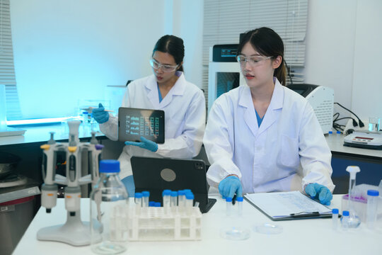Two Asian Female Scientists Working Together in Modern Laboratory with Tablet and Test Tubes