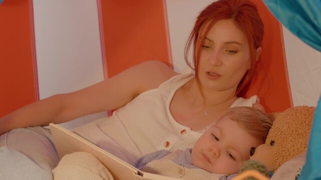 A loving mother shares heartfelt stories with her son in a cozy tent, gently caressing him as he slowly falls asleep next to his teddy bear.