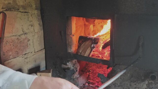Fiery orange flames and glowing red embers in a brick oven. Unidentifiable person using a metal fire poker to move burning logs and coals