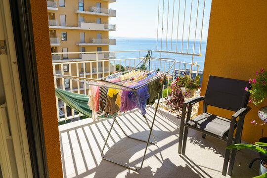 Sunlit apartment balcony overlooking the blue Adriatic Sea, with colorful laundry drying on a folding rack, potted flowers, and outdoor seating