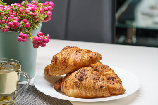 Freshly baked chocolate croissants served on a white plate with a warm beverage and a vibrant pink-berried ornamental plant, photographed in a contemporary home setting