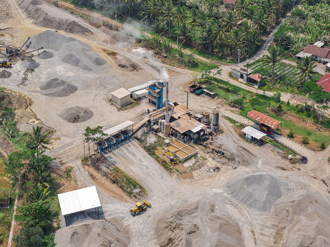 Aerial view of a cement plant with industrial machinery, gravel piles, and smoke rising from a chimney, surrounded by palm trees in Yogyakarta, Special Region of Yogyakarta, Indonesia.