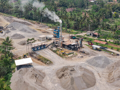Aerial view of a cement plant with smoking chimney, piles of gravel and sand, industrial machinery, and lush tropical vegetation in Yogyakarta, Special Region of Yogyakarta, Indonesia.