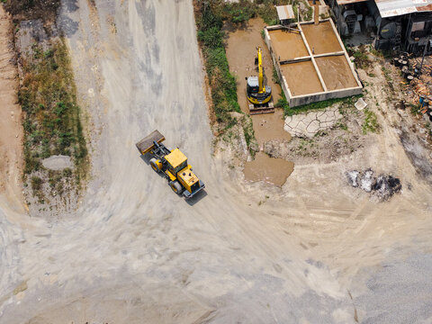 Aerial view of a yellow wheel loader moving on a dirt path near a cement plant with an excavator working in a muddy area in Yogyakarta, Special Region of Yogyakarta, Indonesia.