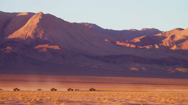 Off-road vehicle convoy drives left to right across a broad desert plain, raising pale dust beneath warm sunlight as rugged layered mountains dominate the distant background