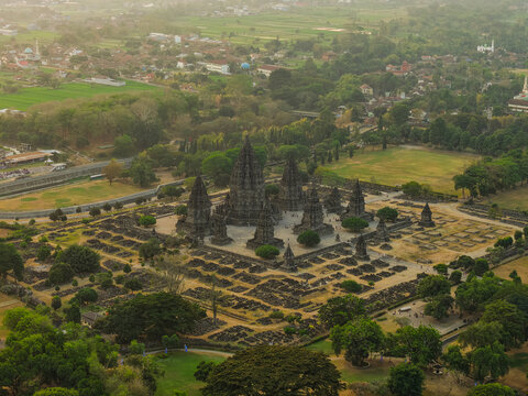 Aerial view of Prambanan Temple with its iconic stone spires, surrounding ruins, lush trees, and grassy grounds under soft golden hour light in Yogyakarta, Special Region of Yog, Indonesia.