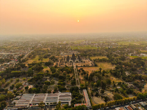 Aerial view of Prambanan Temple complex surrounded by lush greenery and urban landscape during a golden sunset in Yogyakarta, Special Region of Yogyakarta, Indonesia.