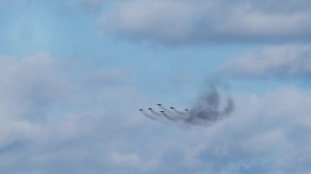 Wide shot captures six combat aircraft cruising horizontally through thick cloudy airspace. Aerobatic squadron releases long dark smoke trails during military flight display.