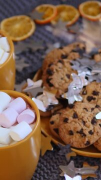 Chocolate chip cookies and marshmallows fill orange dishes as silver star confetti glides through cozy holiday arrangement. Seasonal sweets and citrus accents suggest festive celebration.