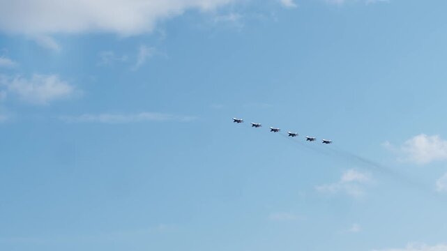 Wide shot captures six military fighter jets flying in straight line formation from distance across bright blue sky with clouds. Squadron maintains precise spacing during flyby.