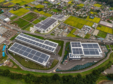 Aerial view of industrial buildings with extensive solar panel arrays on the rooftops, surrounded by lush green rice fields and local infrastructure in Atsugi, Kanagawa, Japan.