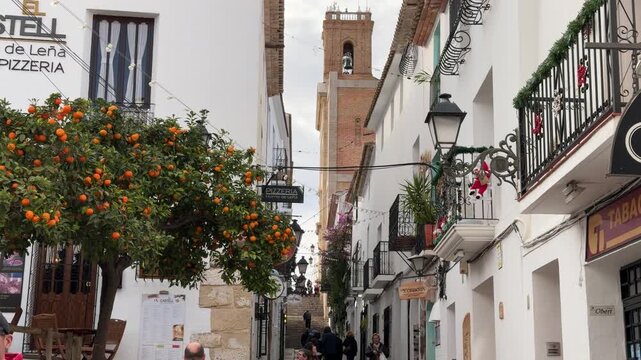 Eye-level shot strolling through a charming cobblestone street featuring an ornate orange tree and a historic brick bell tower in a coastal Mediterranean town of Altea in Spain.