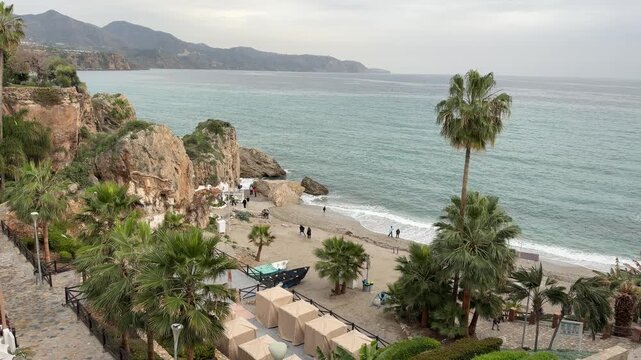 High angle scenic view of a rocky Mediterranean beach with palm trees and sun canopies along the rugged coastal cliffs of Nerja in Malaga, Spain.