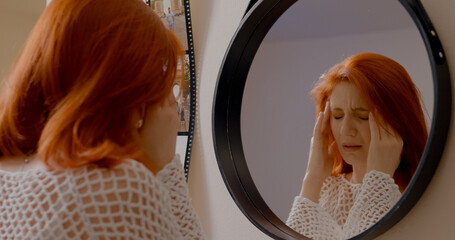 A redhead woman stands in front of her mirror, visibly distressed as she deals with headaches and migraines before heading out of the house. © Binzari