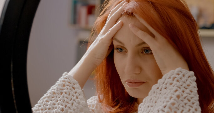 Woman with long red hair feeling strong headache and migraine touching her head and forehead in front of a mirror