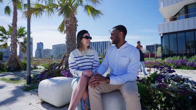 A young Black man and White woman sit together, smile and have a relaxed conversation beside flowers and palm trees.
