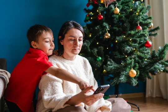 Mother and son use smartphone beside Christmas tree at home