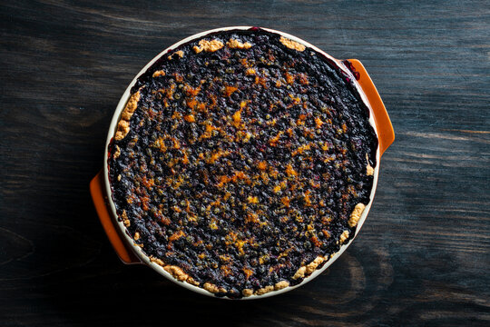 Homemade blueberry pie in baking dish on dark wooden background, top view, closeup. Freshly baked blueberry pie with dark bubbling berry filling and golden crust in ceramic dish on table