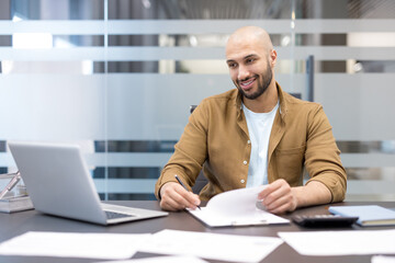 Obraz premium Remote work and business productivity, smiling man working on laptop and reviewing documents in modern office, Professional employee analyzing paperwork and planning strategy at desk
