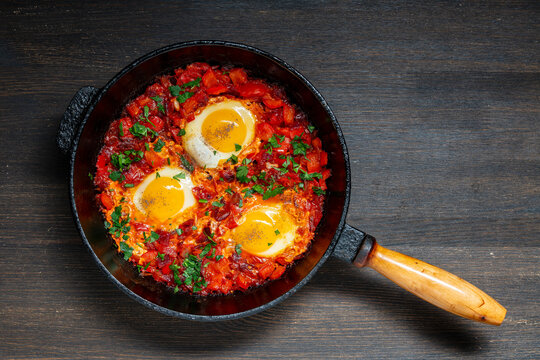 Shakshuka in cast iron skillet, top view, closeup. Traditional middle eastern shakshuka with three fried eggs poached in spicy tomato and pepper sauce with fresh herbs in pan on dark background