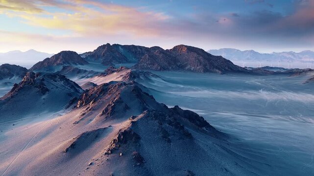 Aerial view of rugged desert mountain ridges bathed in warm sunrise light with soft shadows and pastel clouds in Xinjiang, China.
